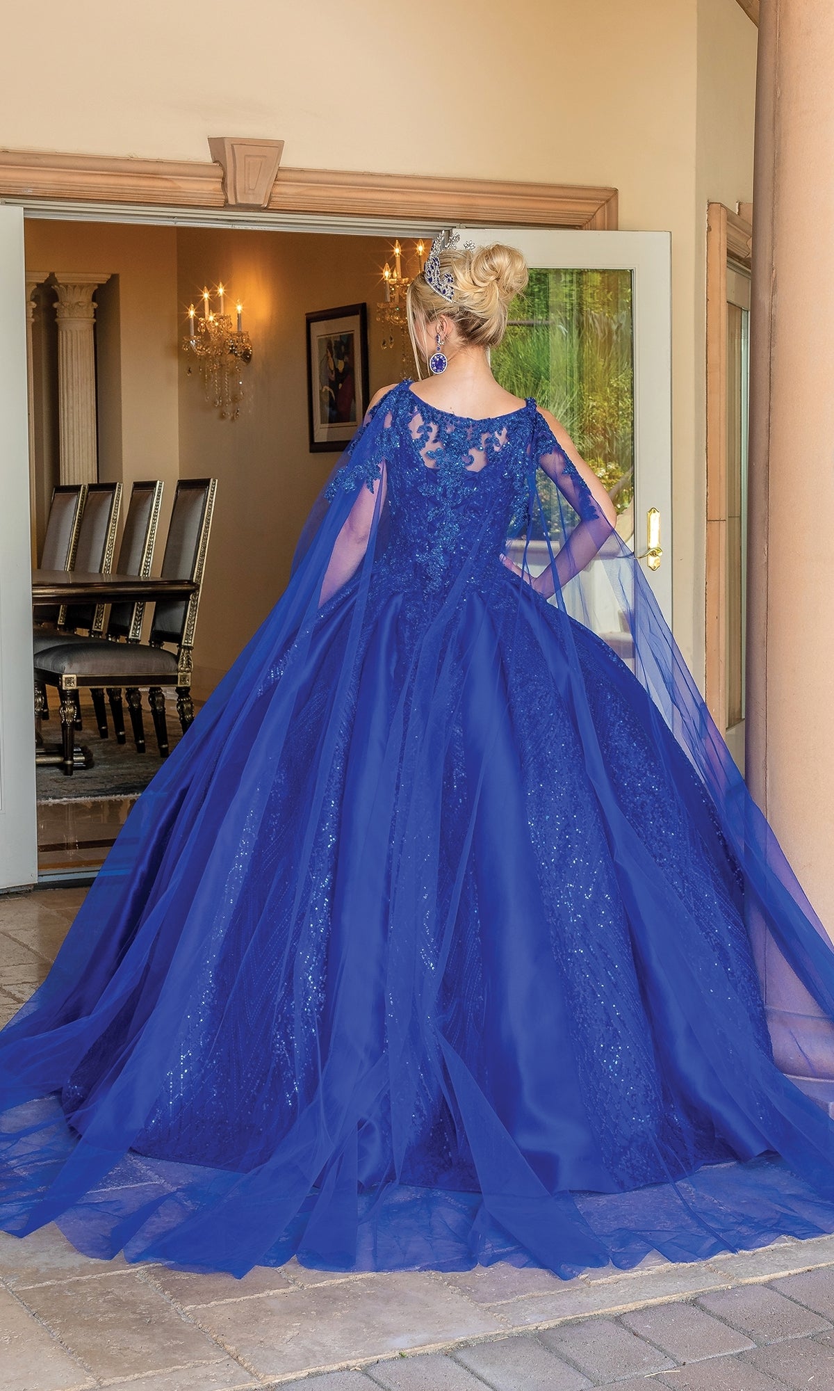 Wearing the Quinceanera Dress 1680 By Dancing Queen, a person in an elaborate blue ball gown with a sweetheart neckline and removable cape stands outside near large doors, with a dining room visible inside.
